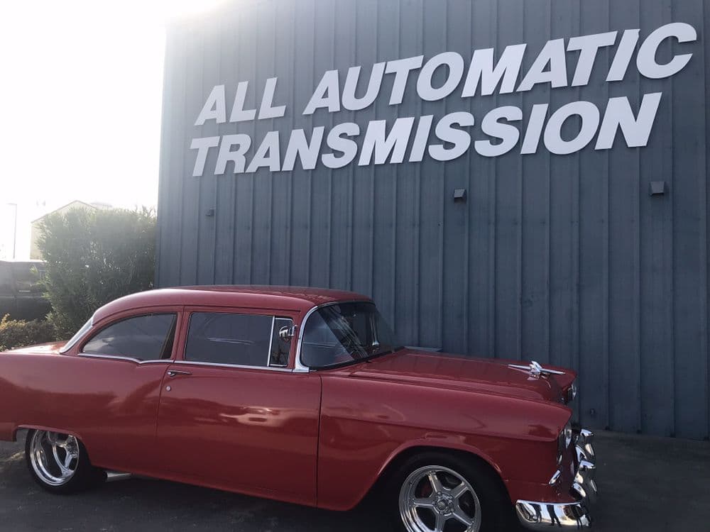 A restored red 1955 Chevrolet parked beneath the All Automatic Transmission wall sign